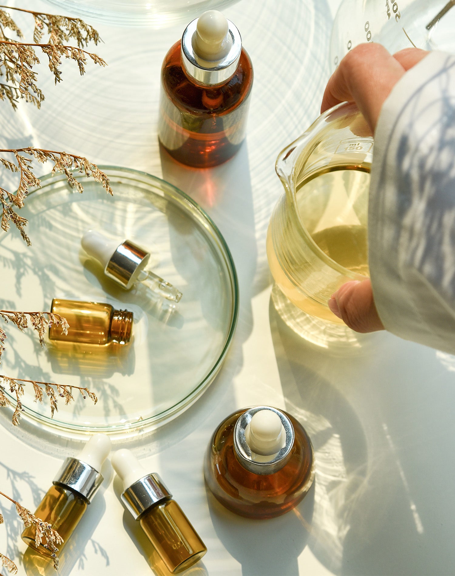 Glass dropper bottle with amber liquid on a white surface with glass containers and dried plants.