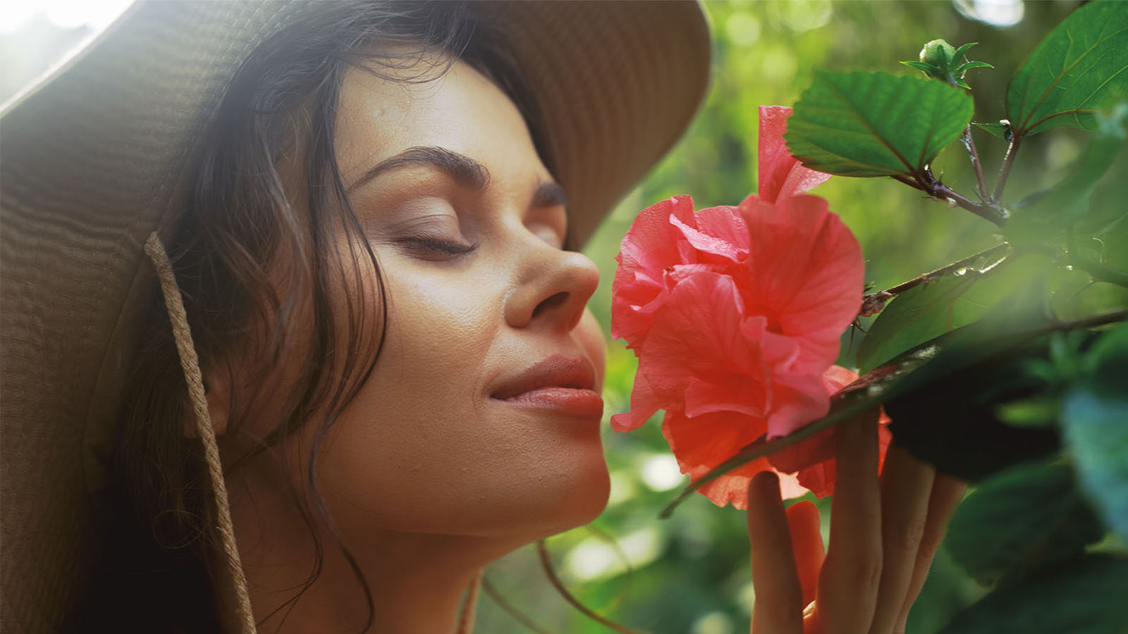 A woman wearing a hat and smelling a flower in the sun in a park or garden.
