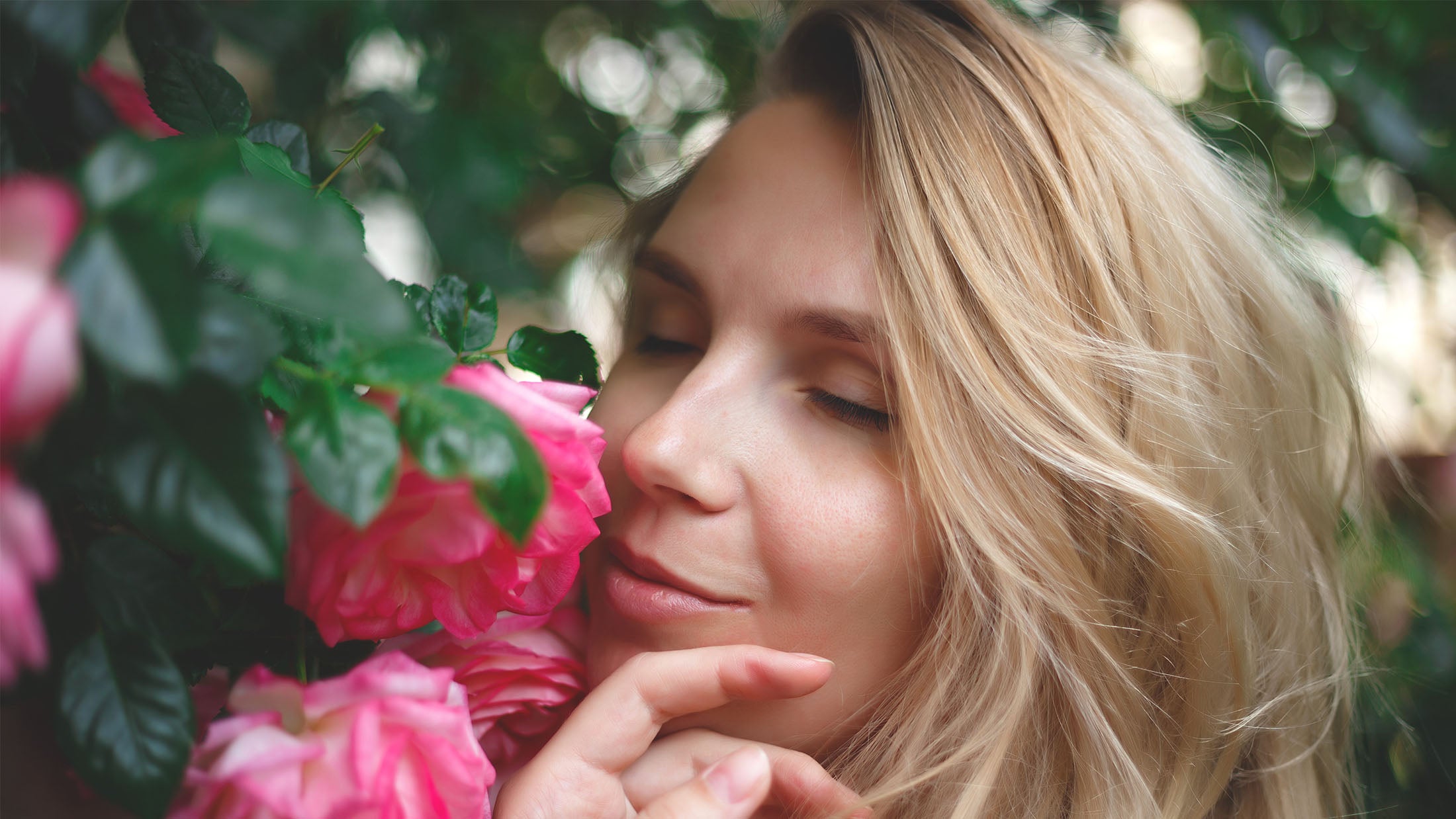Woman with blonde hair smelling pink flowers among green leaves.